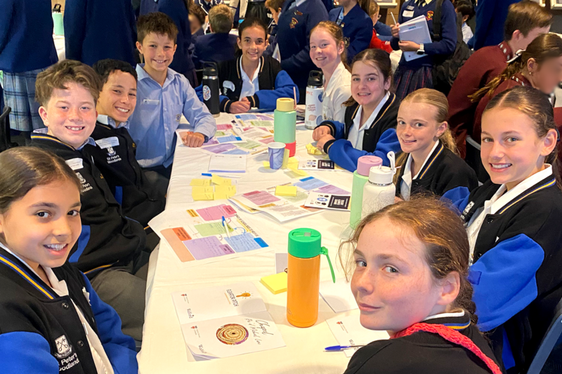 A group of St Peter's Woodlands student leaders smiling and gathered around a table at the Anglican Schools Network Leadership Workshop. The table is filled with colorful worksheets, documents, and water bottles, as the students collaborate on leadership activities.