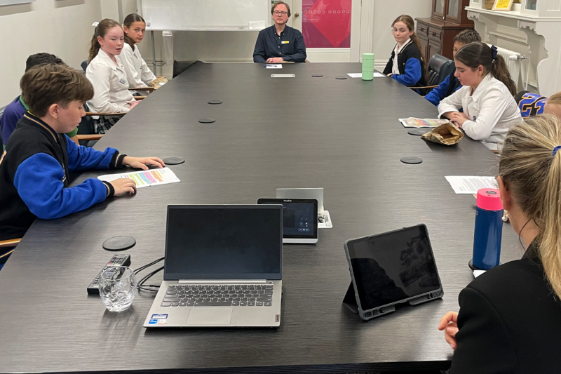 Students presenting their leadership ideas at a roundtable meeting with the Business Manager. The group is gathered around a large table, discussing solutions for school accessibility, with laptops and documents visible in front of them.